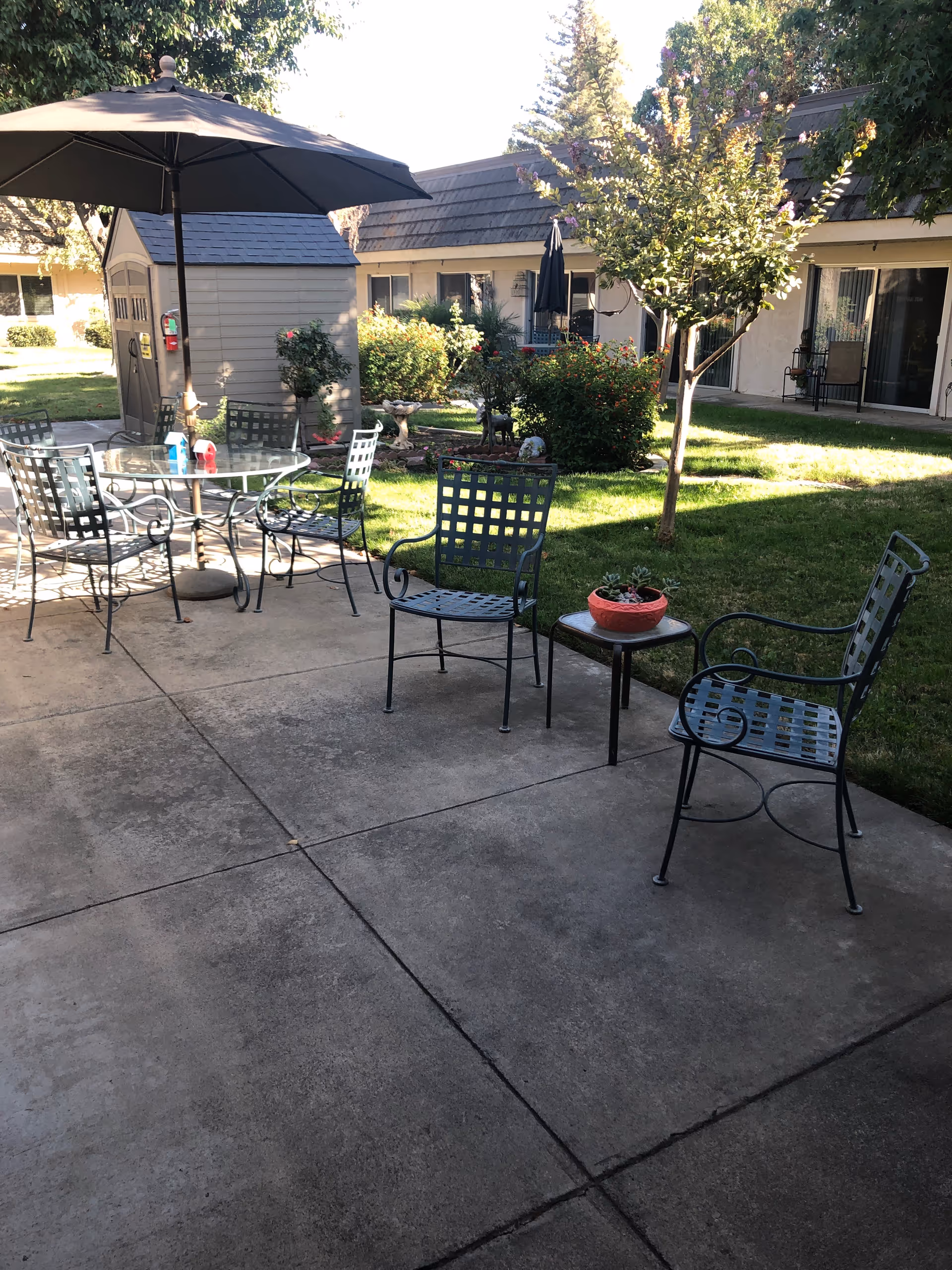 Outdoor patio with metal chairs, table, umbrella, and greenery at Cypress Assisted Living.