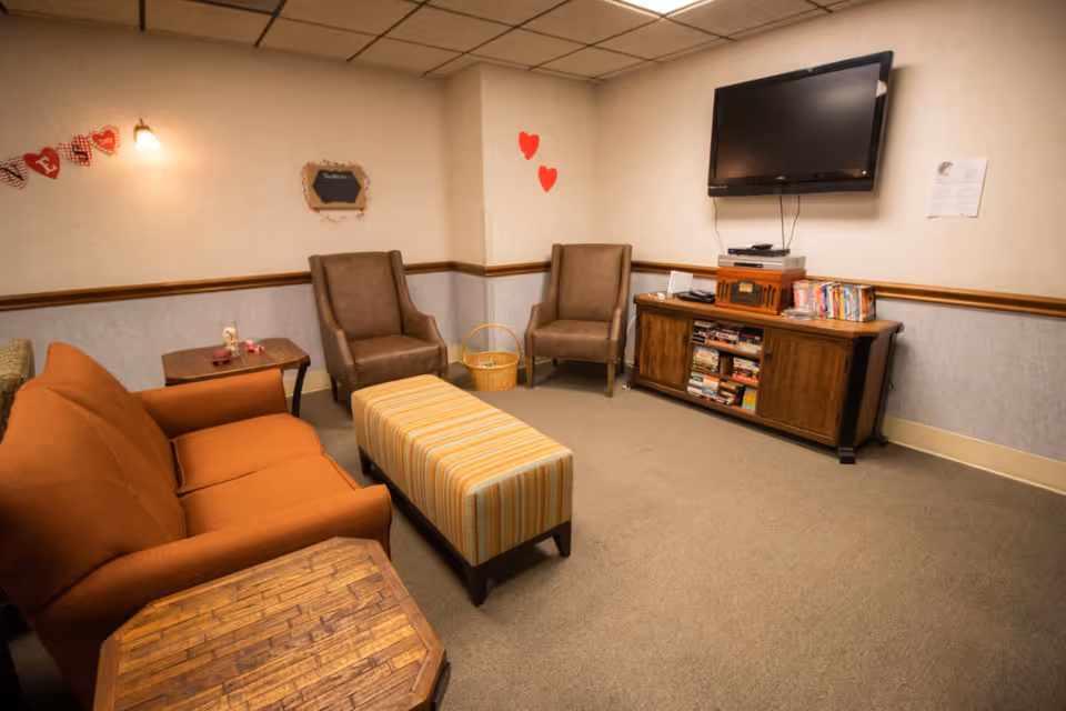 Small common living room with an orange sofa, two armchairs, a striped ottoman, wall-mounted TV, and a media cabinet with books.