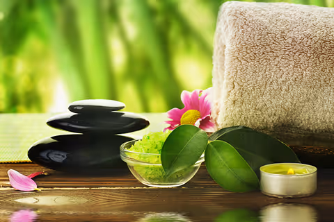 Spa arrangement on a wooden surface: rolled towel, stones, green bath salts, leaves, candle.