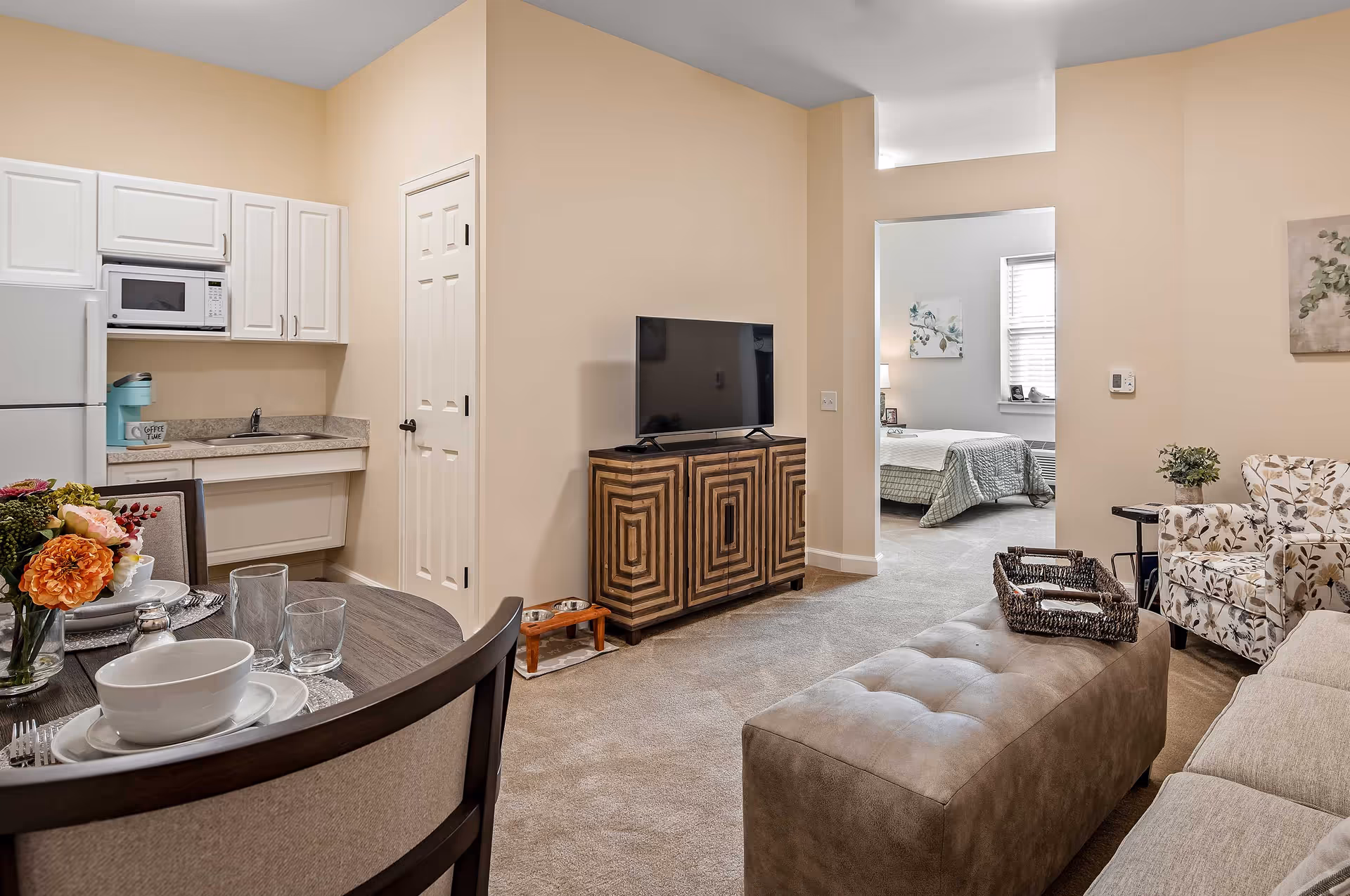 Open-concept living area with a small kitchenette and dining table, TV on a patterned cabinet, seating, and a doorway leading to a bedroom.