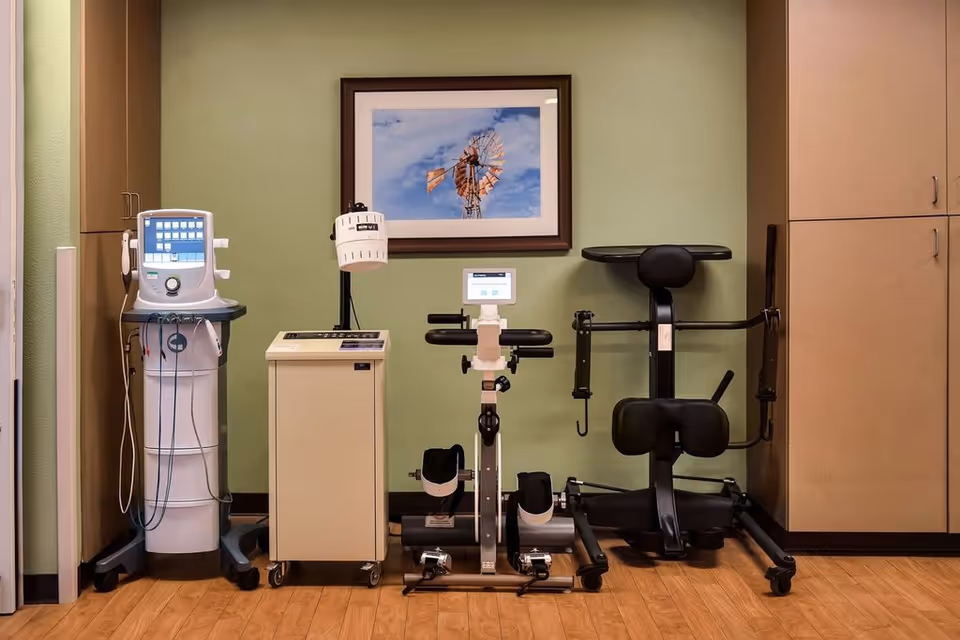 A room with medical and exercise equipment including a white medical device with wires, a beige machine on wheels, a stationary exercise bike, and a black exercise machine. The walls are light green with wooden cabinets on either side and a framed picture of a windmill on the wall.