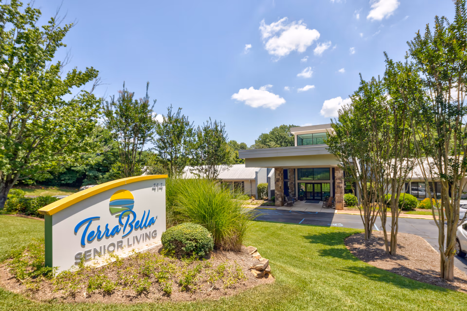 Exterior view of TerraBella Senior Living facility with a clear blue sky, green trees, and well-maintained landscaping. The building entrance is visible with a covered drop-off area and large windows.