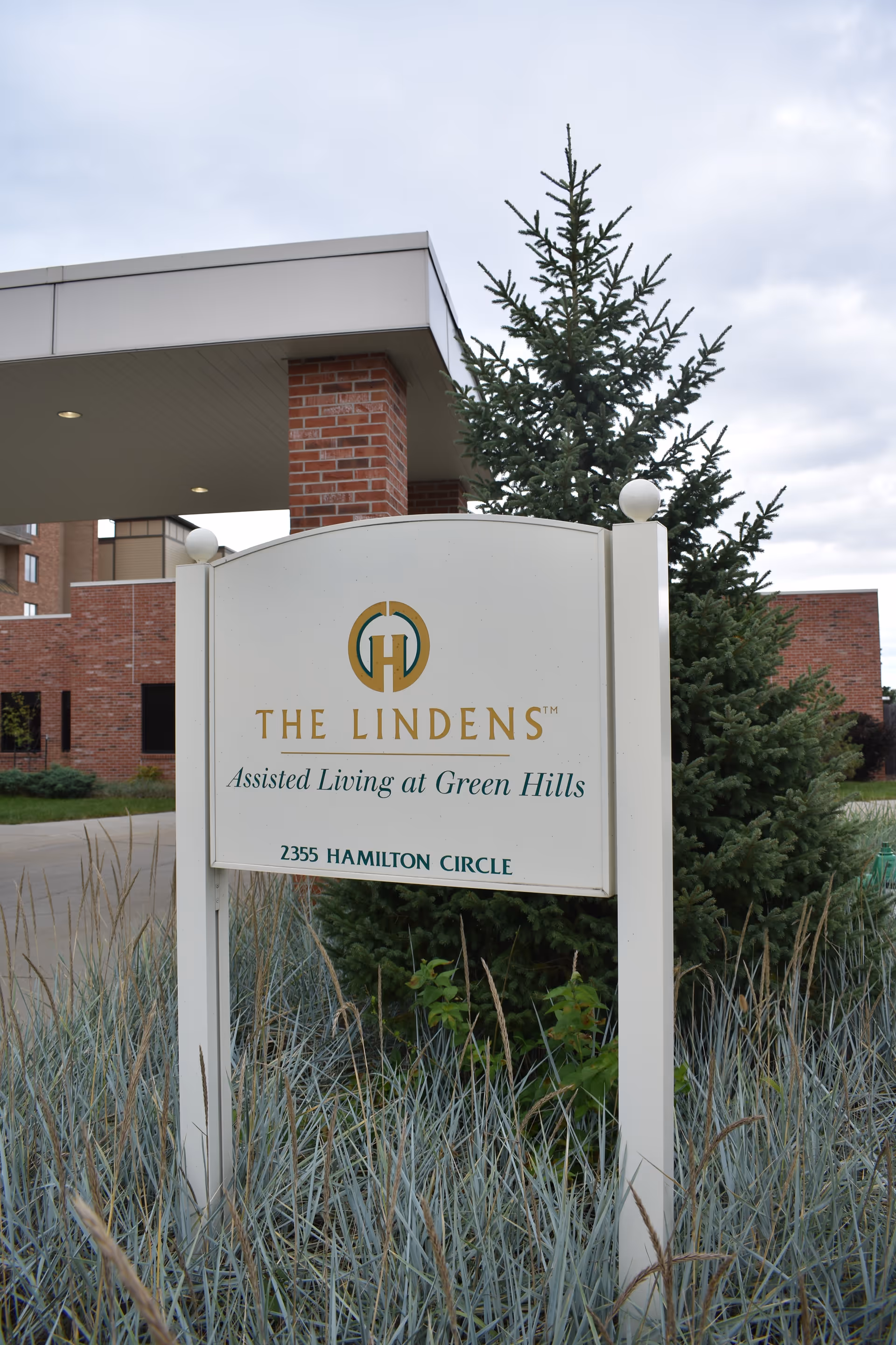A white sign reading 'The Lindens Assisted Living at Green Hills' stands in front of a brick building and evergreen tree.