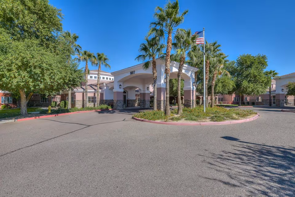 Front exterior view of Chaparral Winds Assisted Living facility with a covered entrance, palm trees, an American flag, and clear blue sky.