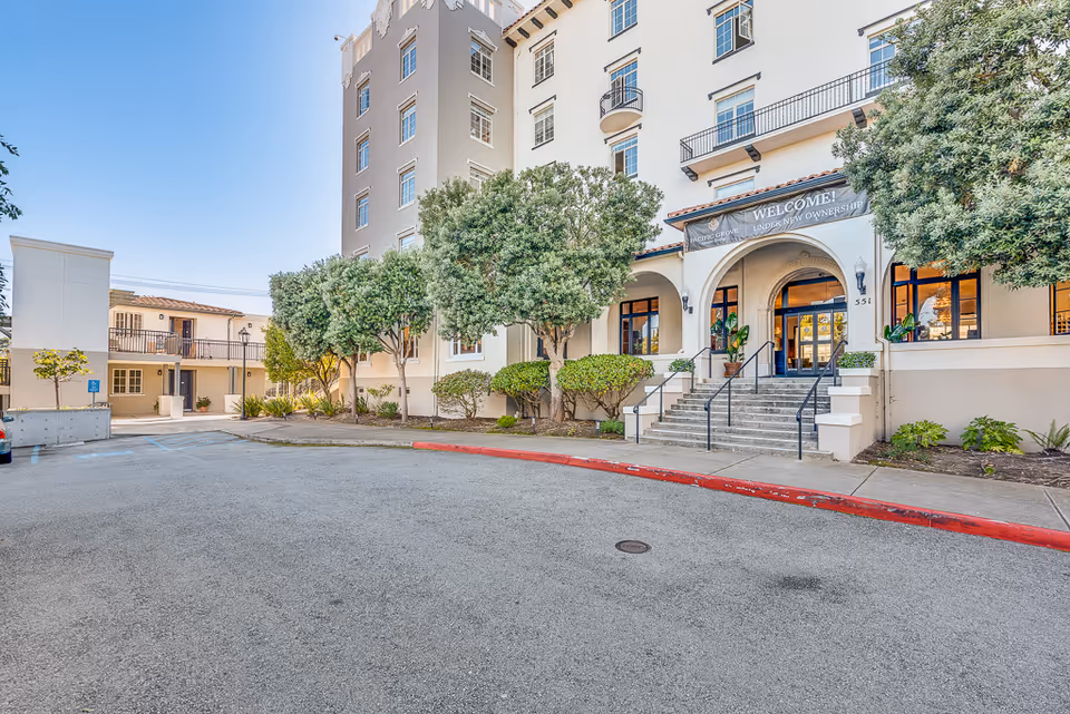 Front exterior of a multi-story senior living building showing steps up to an arched main entrance, trees, and an empty driveway.