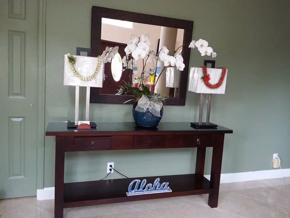 A dark wooden console table against a light green wall with a large rectangular mirror above it. On the table are two lamps with white shades, each decorated with a lei, and a blue vase with white orchids and greenery. Below the table is a decorative sign that says 'Aloha'.