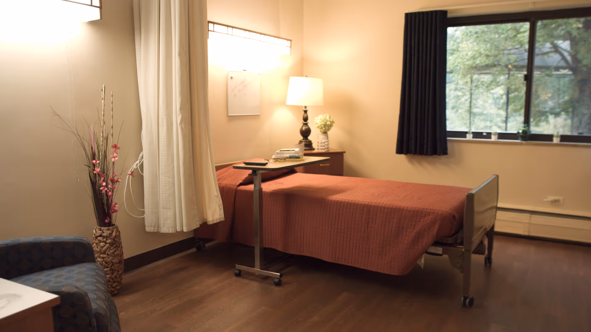 A simple, cozy bedroom in a senior living facility with a single bed covered in a rust-colored blanket, a small overbed table with books and a remote, a bedside table with a lamp and a flower vase, a blue patterned armchair, a tall decorative vase with flowers, and a window with dark curtains showing greenery outside.
