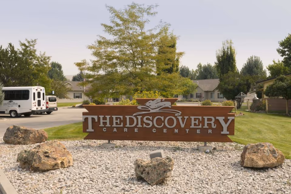 Outdoor view of the Discovery Care Center sign made of wood with silver letters, surrounded by rocks and landscaping. In the background, there are trees, a parking area with a white shuttle bus, and residential-style buildings.