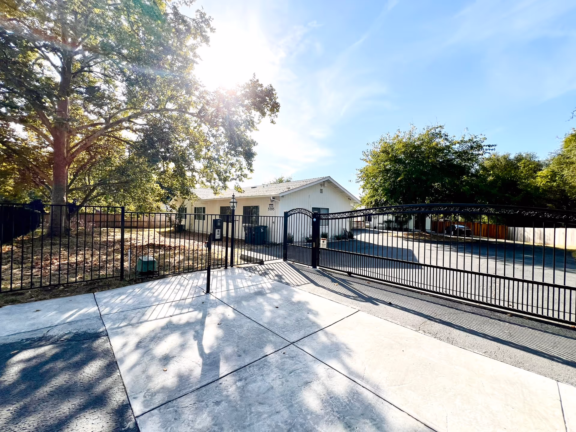 Sunlit entrance gate to a senior living facility with a paved driveway, black metal fencing, and a white building in the background surrounded by trees.