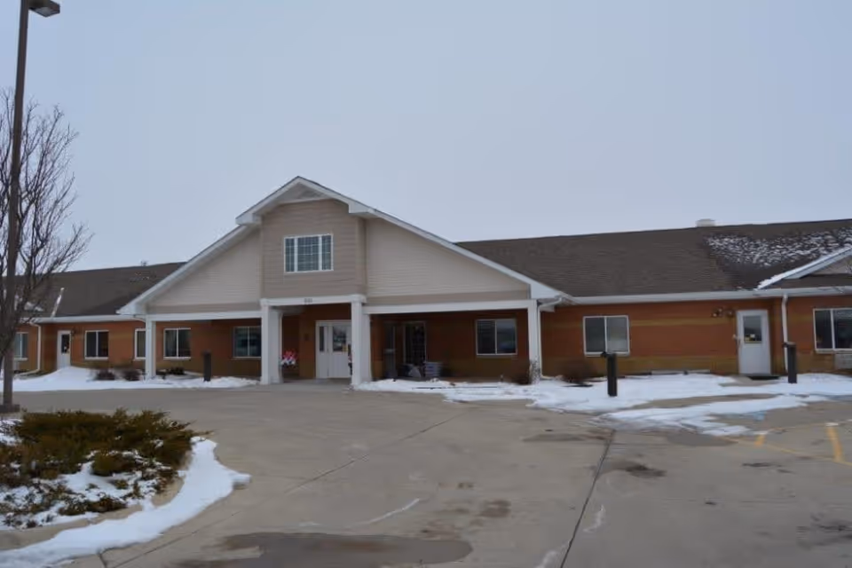 Front exterior view of a single-story senior living facility building with a covered entrance supported by white columns. There is some snow on the ground and patches of bare pavement in the parking area. The building has a combination of brick and siding with multiple windows and doors.