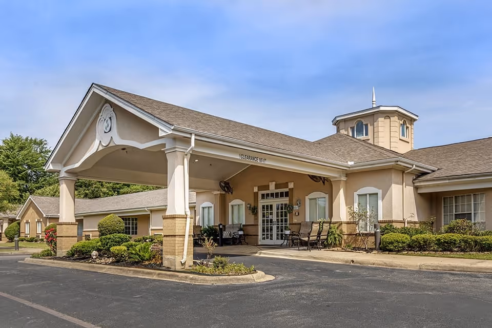 Exterior view of a single-story senior living facility building with a covered entrance, beige walls, and a small tower-like structure on the roof. There are shrubs and plants around the building and a paved driveway in front.