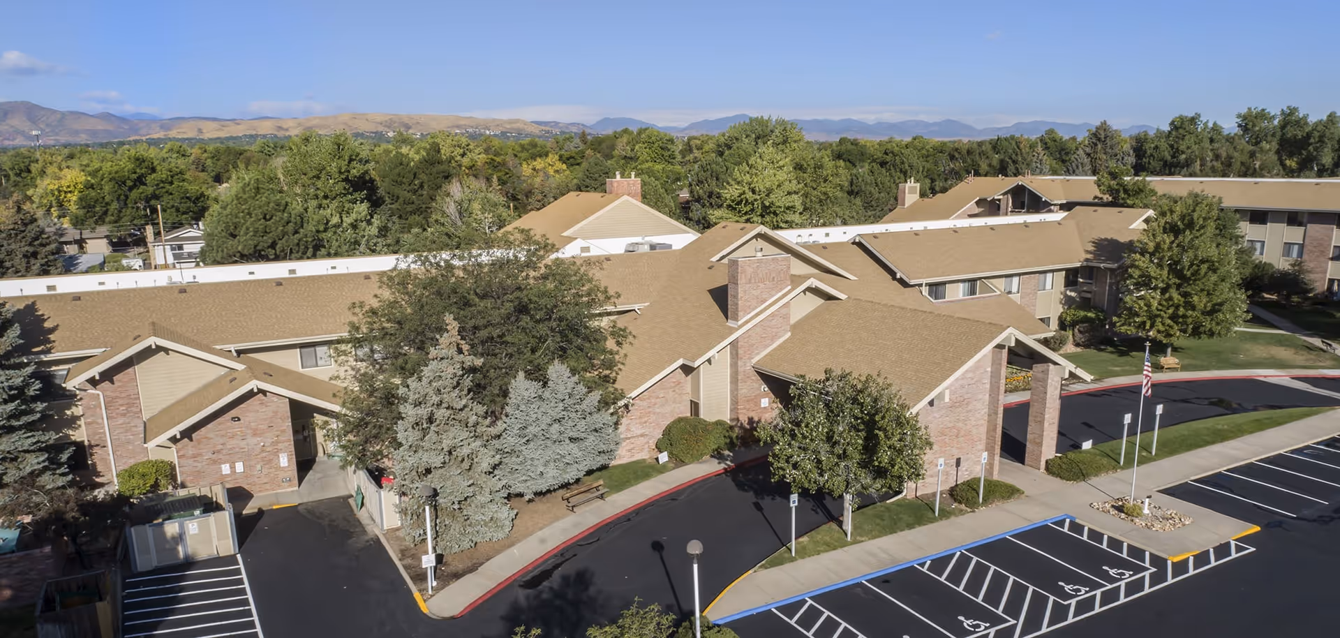 Aerial view of Brookdale Meridian Lakewood senior living facility showing multiple connected buildings with tan roofs and brick walls, surrounded by trees and greenery, with a parking lot featuring several handicap parking spaces and an American flag on a flagpole.