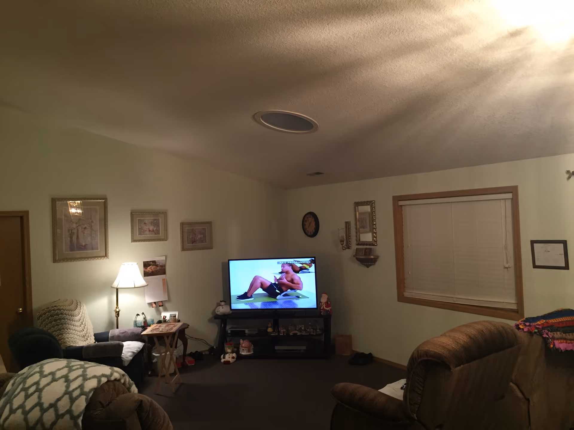 Dim living room with TV, sofa, chairs, lamp, framed pictures, and window blinds.