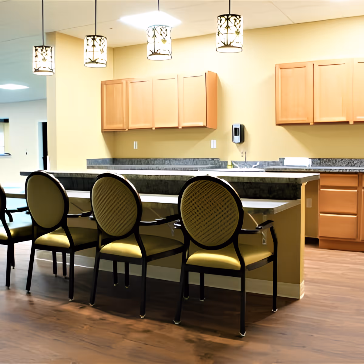 Interior view of a senior living facility kitchen area with a long counter and four cushioned chairs with rounded backs. The kitchen has wooden cabinets mounted on the wall, a granite countertop, and three decorative pendant lights hanging from the ceiling. The floor is wooden, and there is a soap dispenser mounted on the wall near the sink.