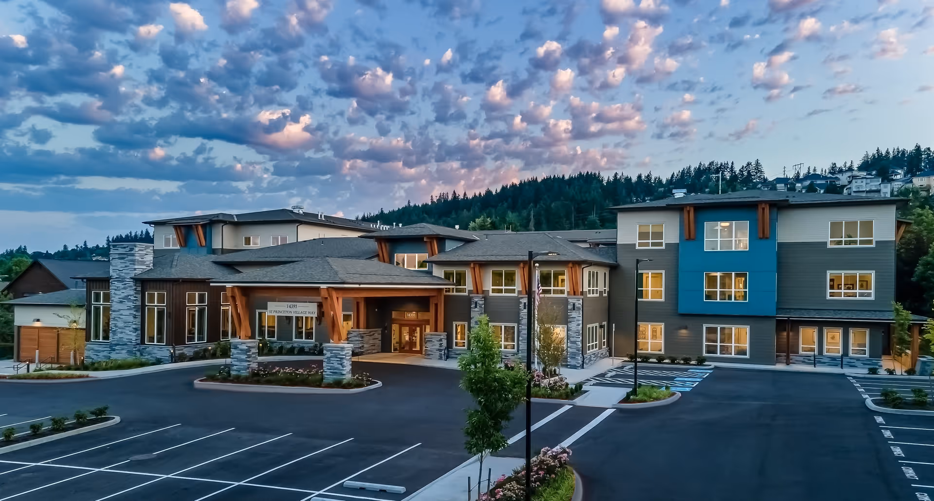Exterior view of a modern assisted living and memory care facility building during dusk with a partly cloudy sky. The building has multiple windows, stone and wood architectural accents, and a covered entrance. The parking lot in front is mostly empty with marked parking spaces and landscaped areas.