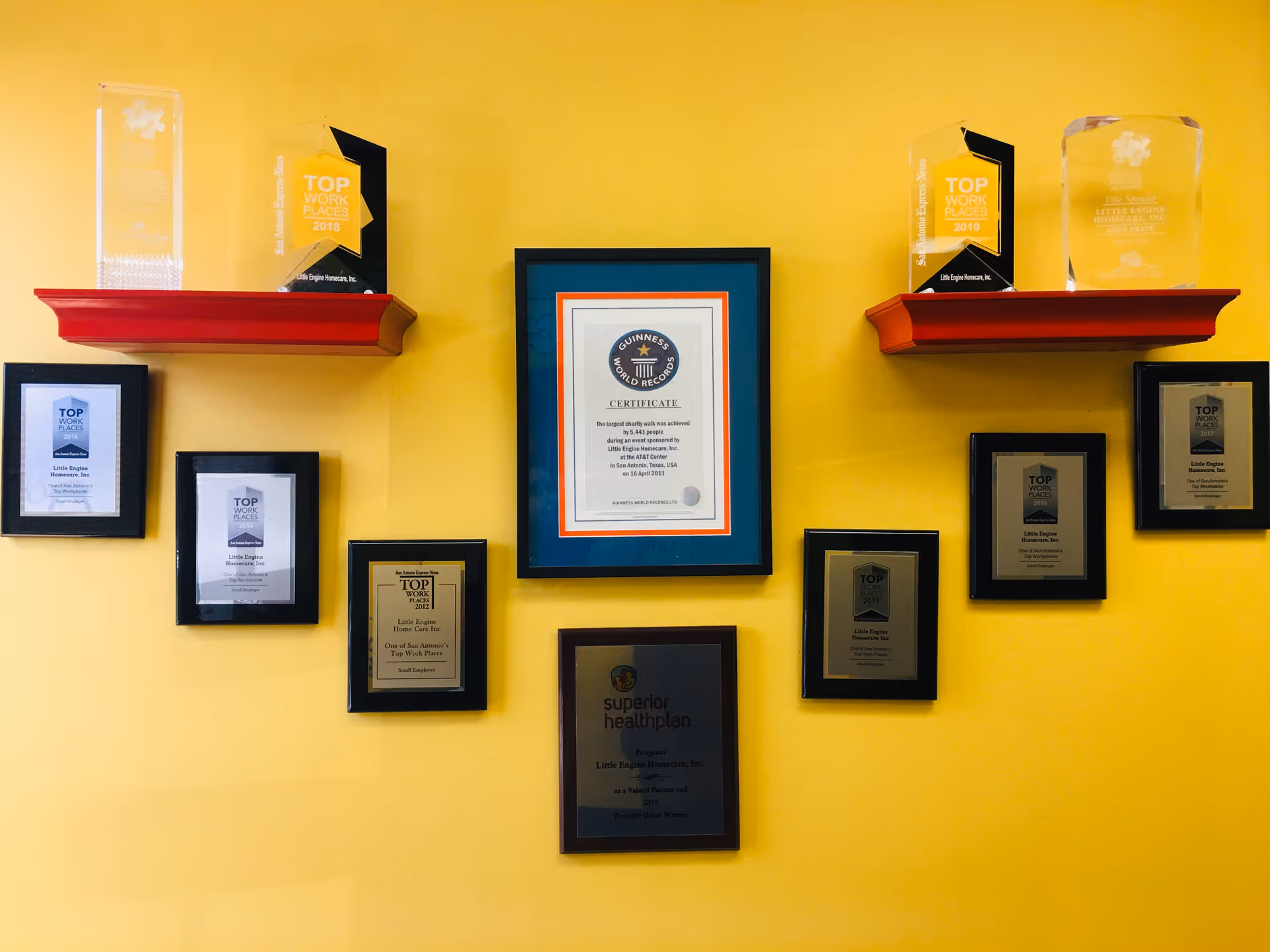 Bright yellow wall with framed certificates and glass awards displayed on red shelves.
