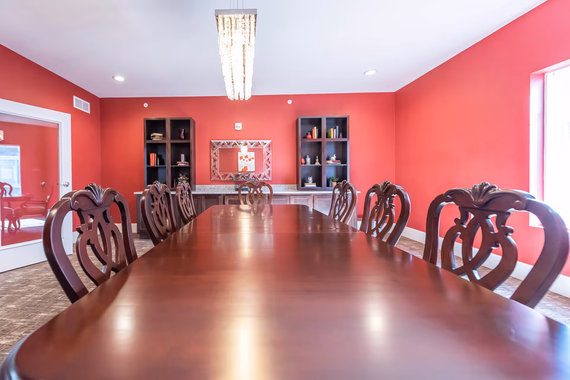Long polished wooden dining table surrounded by ornate chairs in a bright red-walled dining room with shelves, a mirror, and a chandelier.