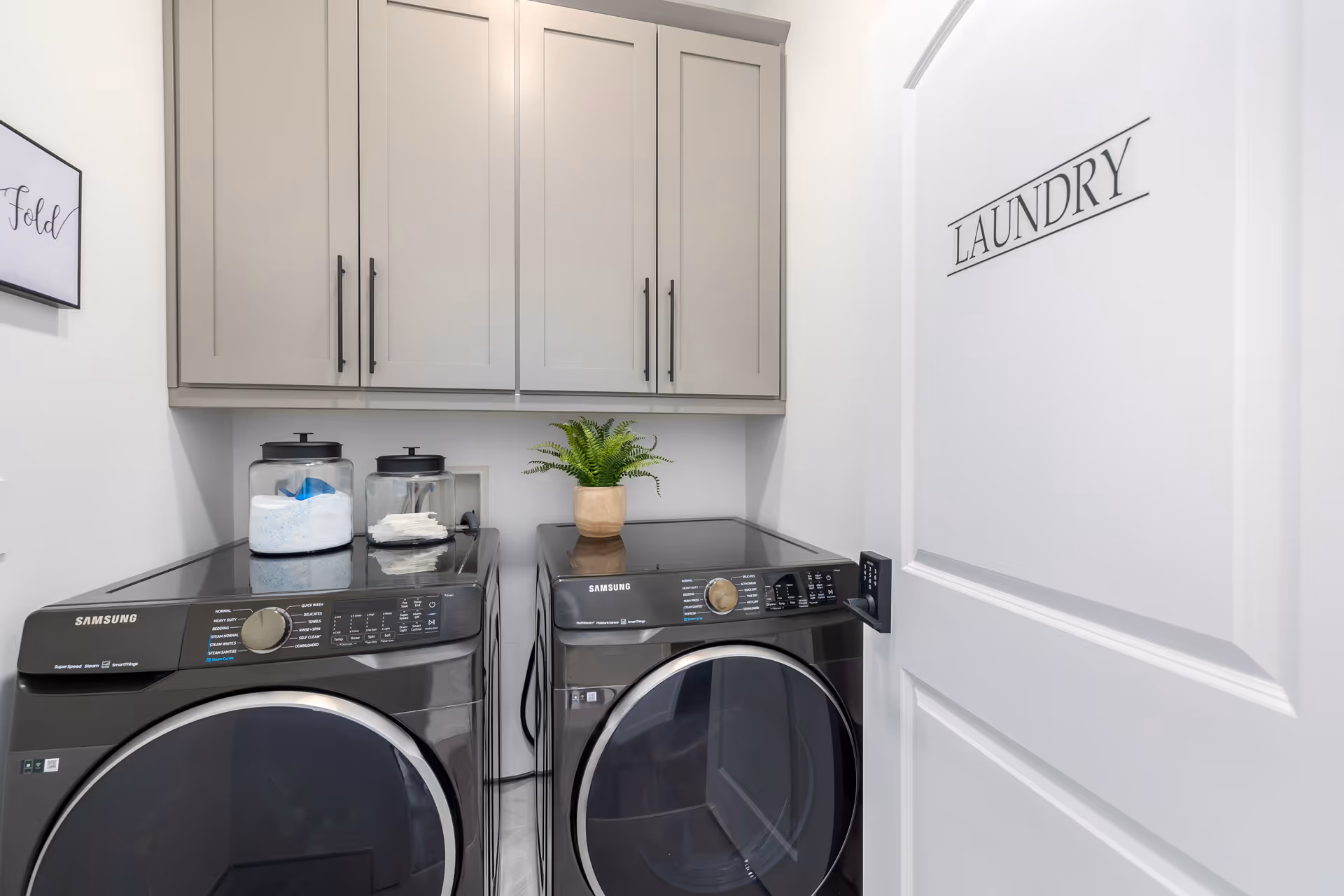 A compact laundry room with two front-loading washers, cabinets, a plant, and a labeled door.