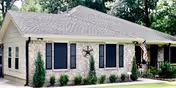 Single-story brick building with a gray shingled roof, black window shutters, and an American flag mounted near the entrance, surrounded by greenery and trees.