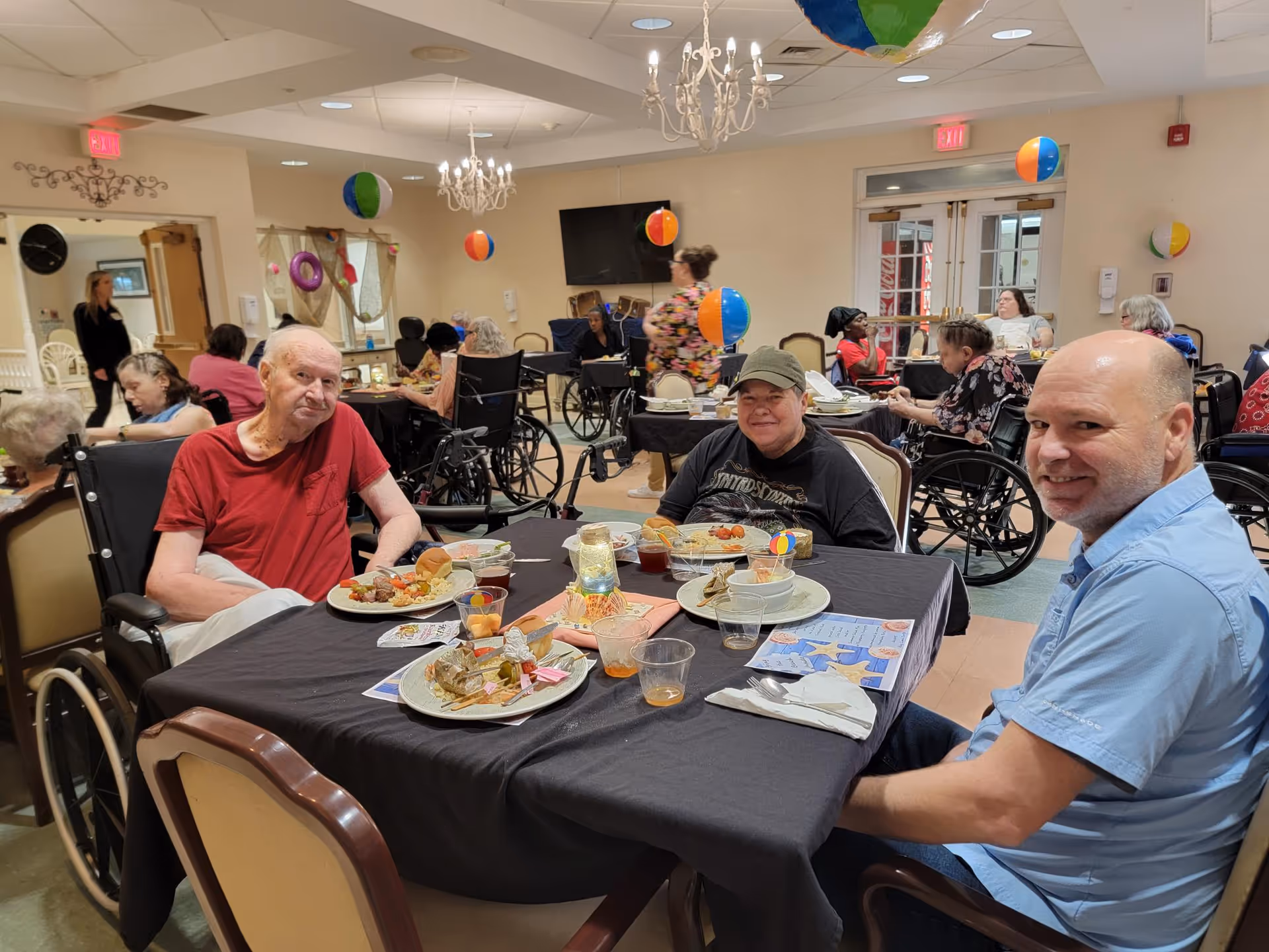 A group of elderly people and a man sitting around a table in a dining room at Bonifay Nursing and Rehab Center. The table is set with plates of food, drinks, and utensils. The room is decorated with colorful beach balls hanging from the ceiling and has chandeliers. Other residents and staff are visible in the background, some in wheelchairs.