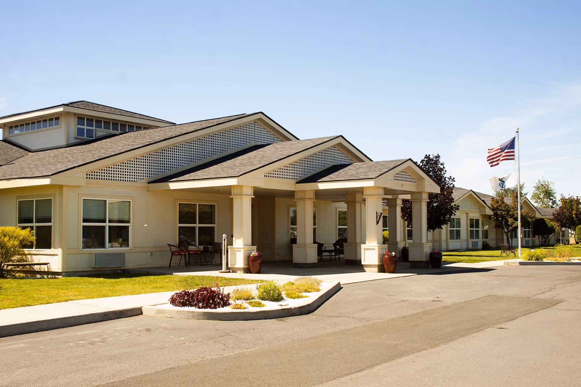 Exterior view of Settler's Park Senior Living facility showing a single-story building with a covered entrance supported by columns, surrounded by a well-maintained lawn, small shrubs, and an American flag on a flagpole under a clear blue sky.