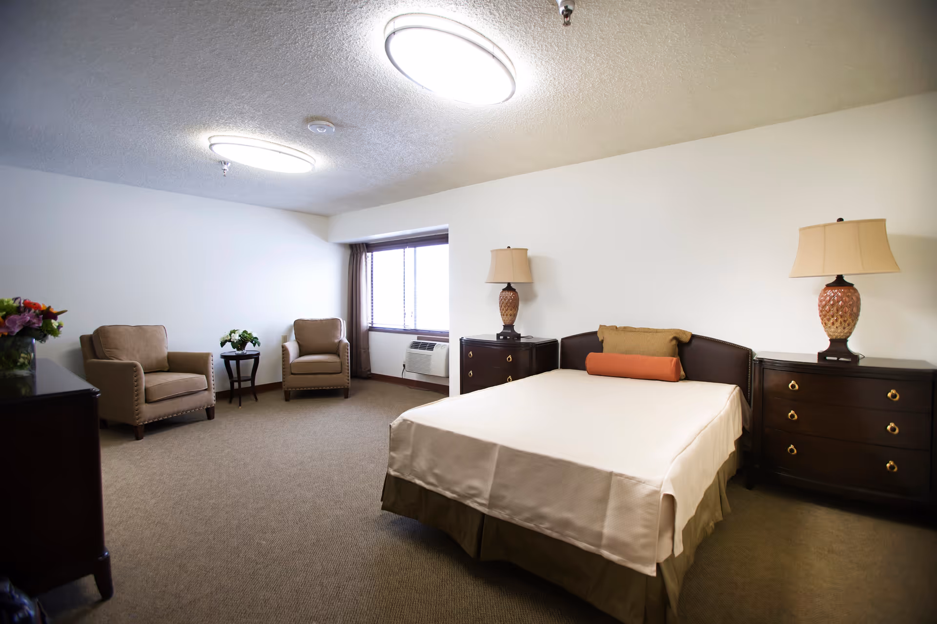 A spacious bedroom with a neatly made bed featuring two pillows and a bolster. On either side of the bed are dark wooden nightstands with matching lamps. Two beige armchairs and a small round table with a flower arrangement are positioned near a window with curtains. The room has beige carpeting and white walls with two ceiling lights.