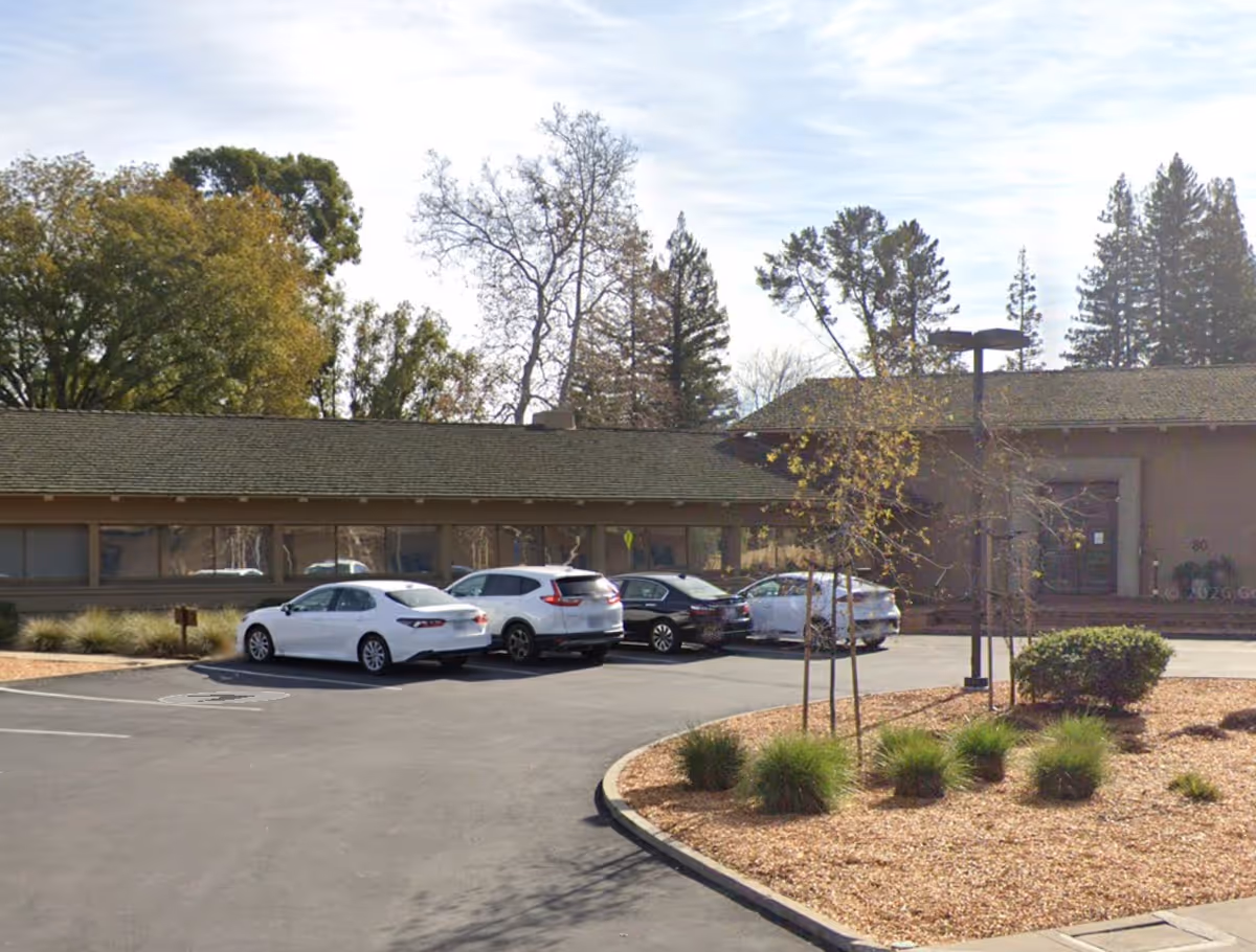 Single-story facility exterior with parked cars, shrubs, and trees in a sunny lot