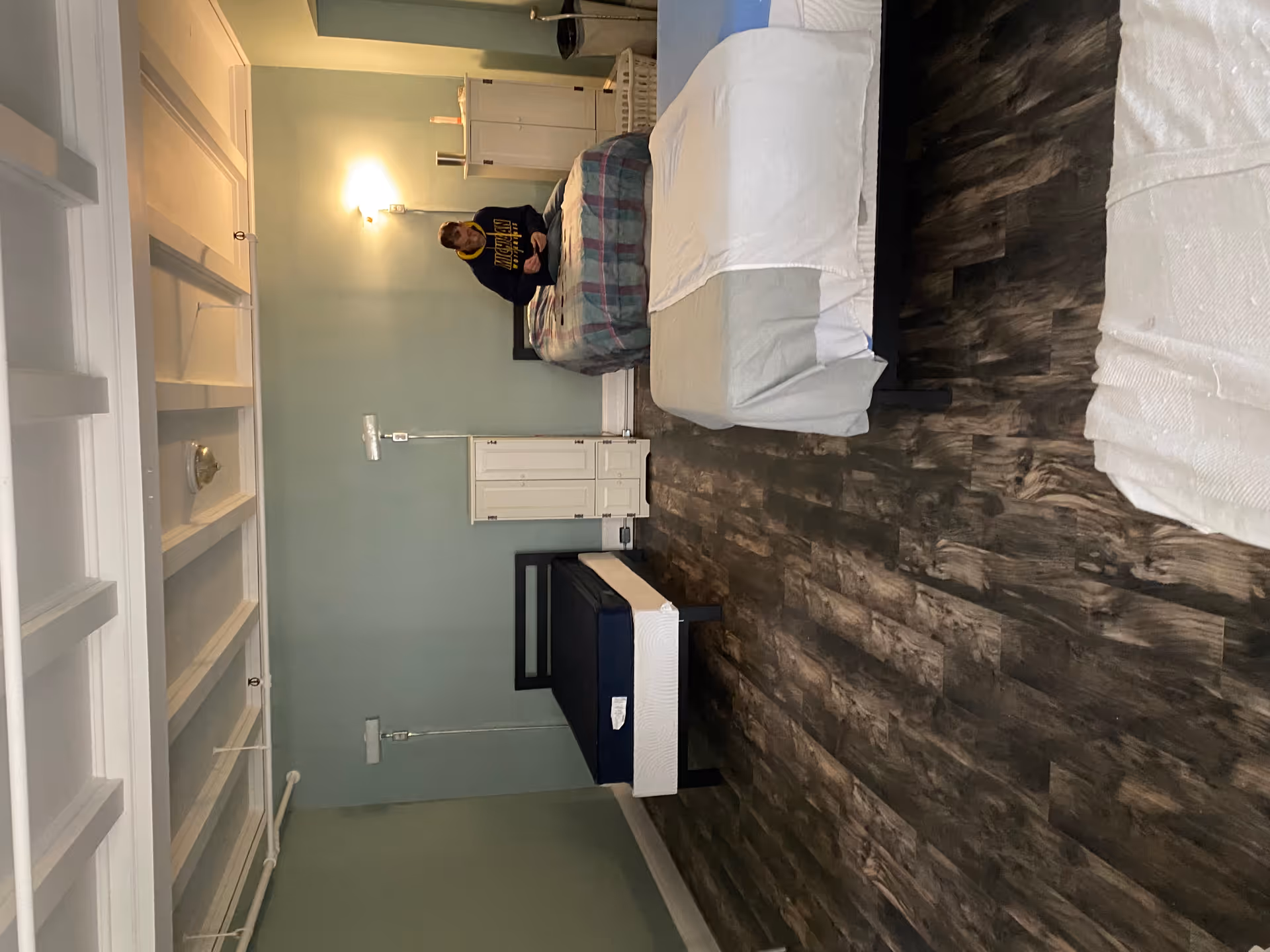 A bedroom in a senior living facility with a bed, dresser, and a person standing nearby.