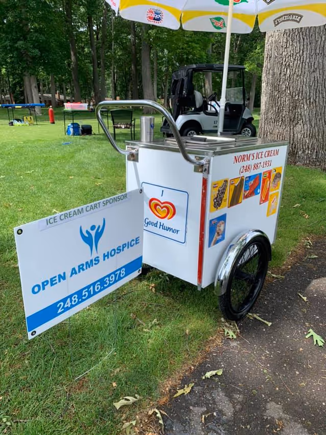 Outdoor park with an Open Arms Hospice ice cream cart beside trees and a golf cart