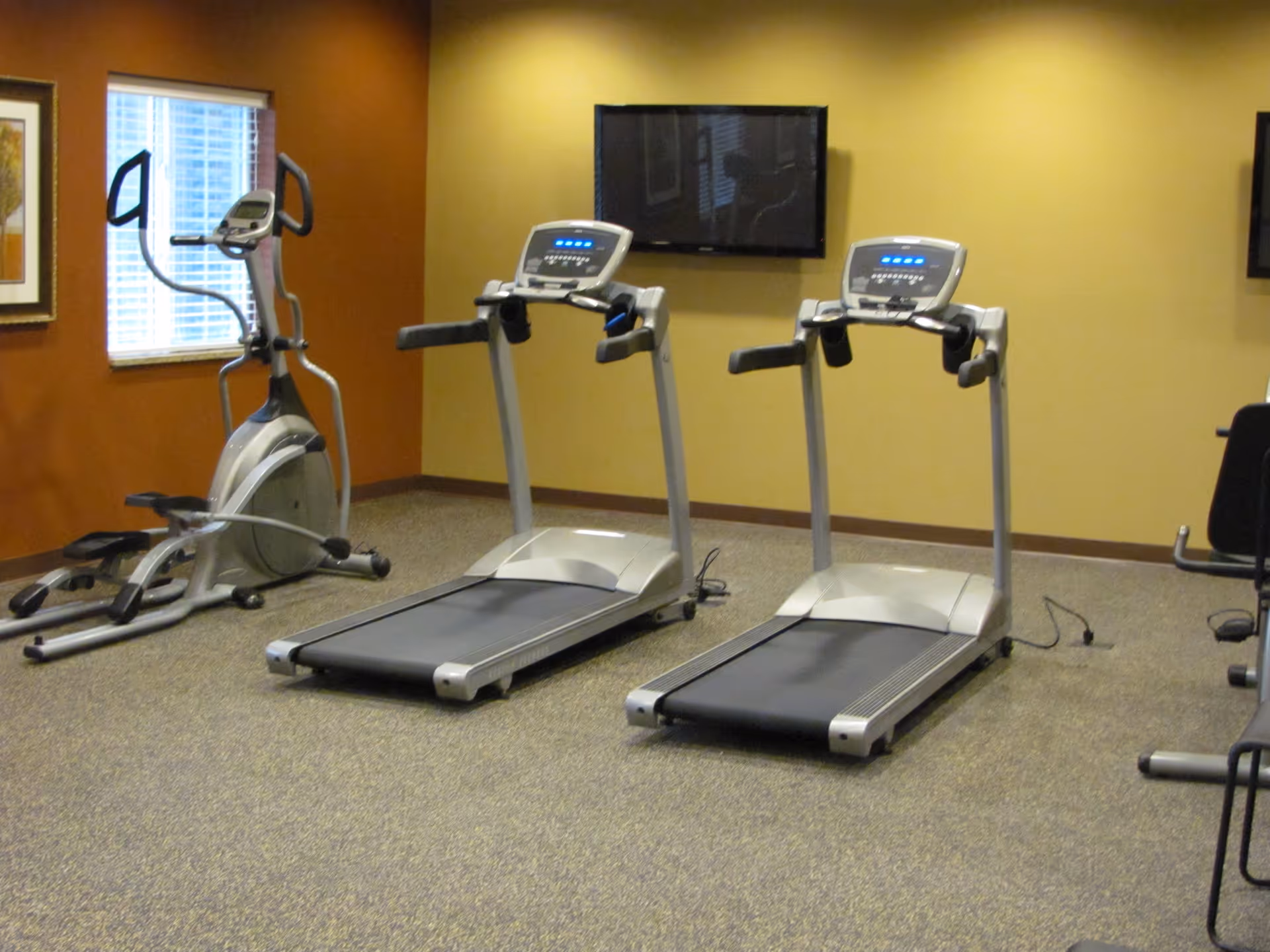 Indoor fitness room with two treadmills, an elliptical machine, a wall-mounted TV, and a window with blinds. The walls are painted in warm tones of yellow and brown.
