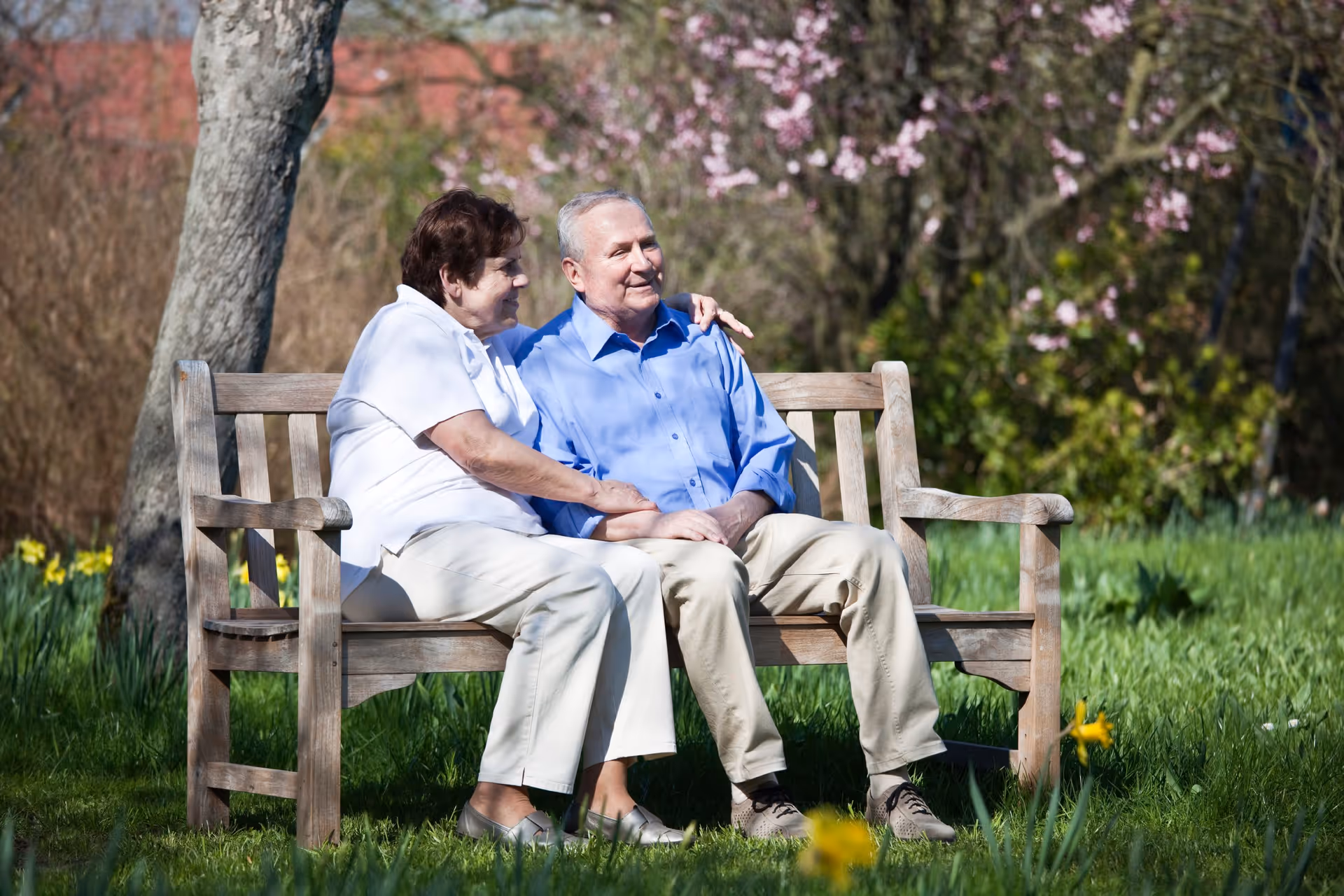 An elderly couple sitting closely together on a wooden bench in a garden. The woman has her arm around the man, and both are smiling. The background features green grass, blooming flowers, and trees with pink blossoms.