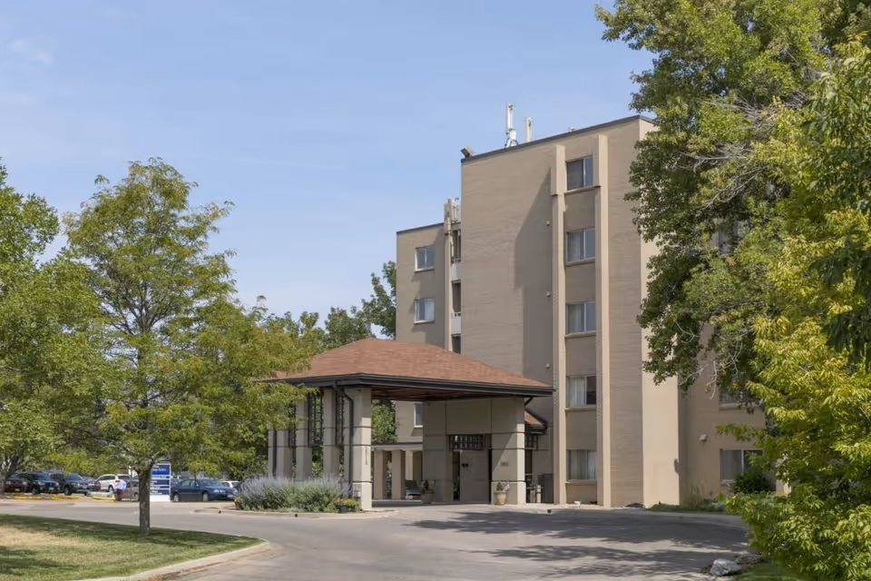 Exterior view of a multi-story beige senior living building with a covered entrance/porte-cochere, surrounded by trees and a parking area.