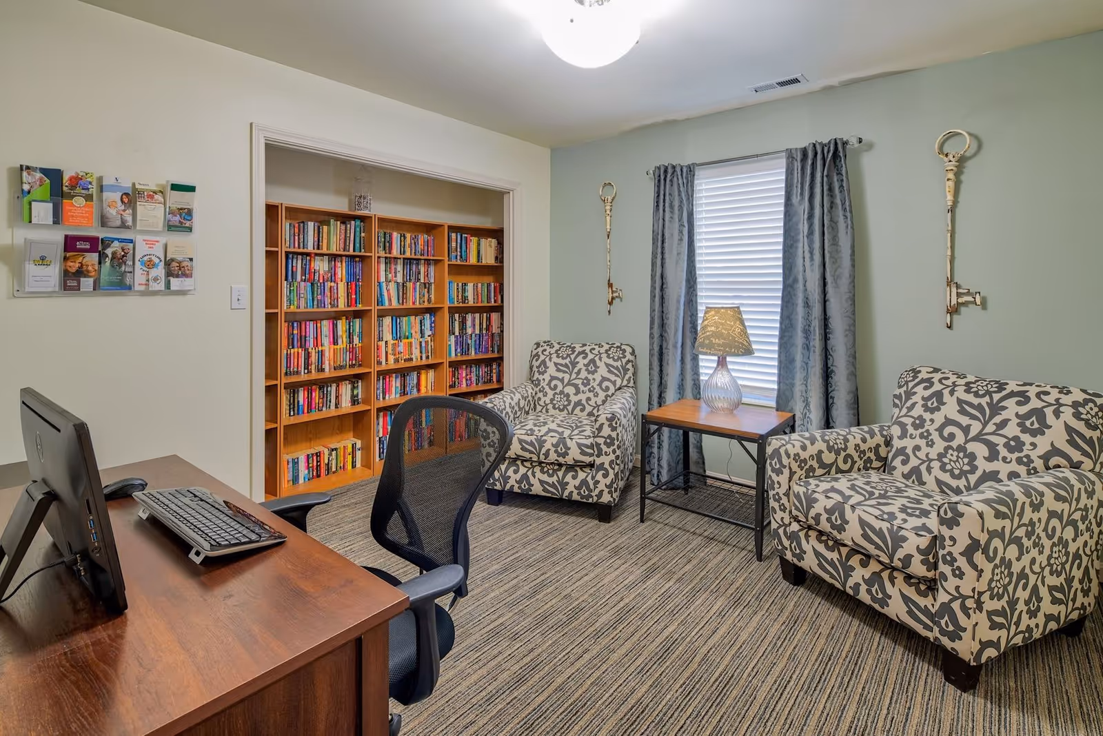 Cozy seating area with two patterned armchairs, a side table and lamp, a bookshelf, and a desk with a computer in a senior living facility room.