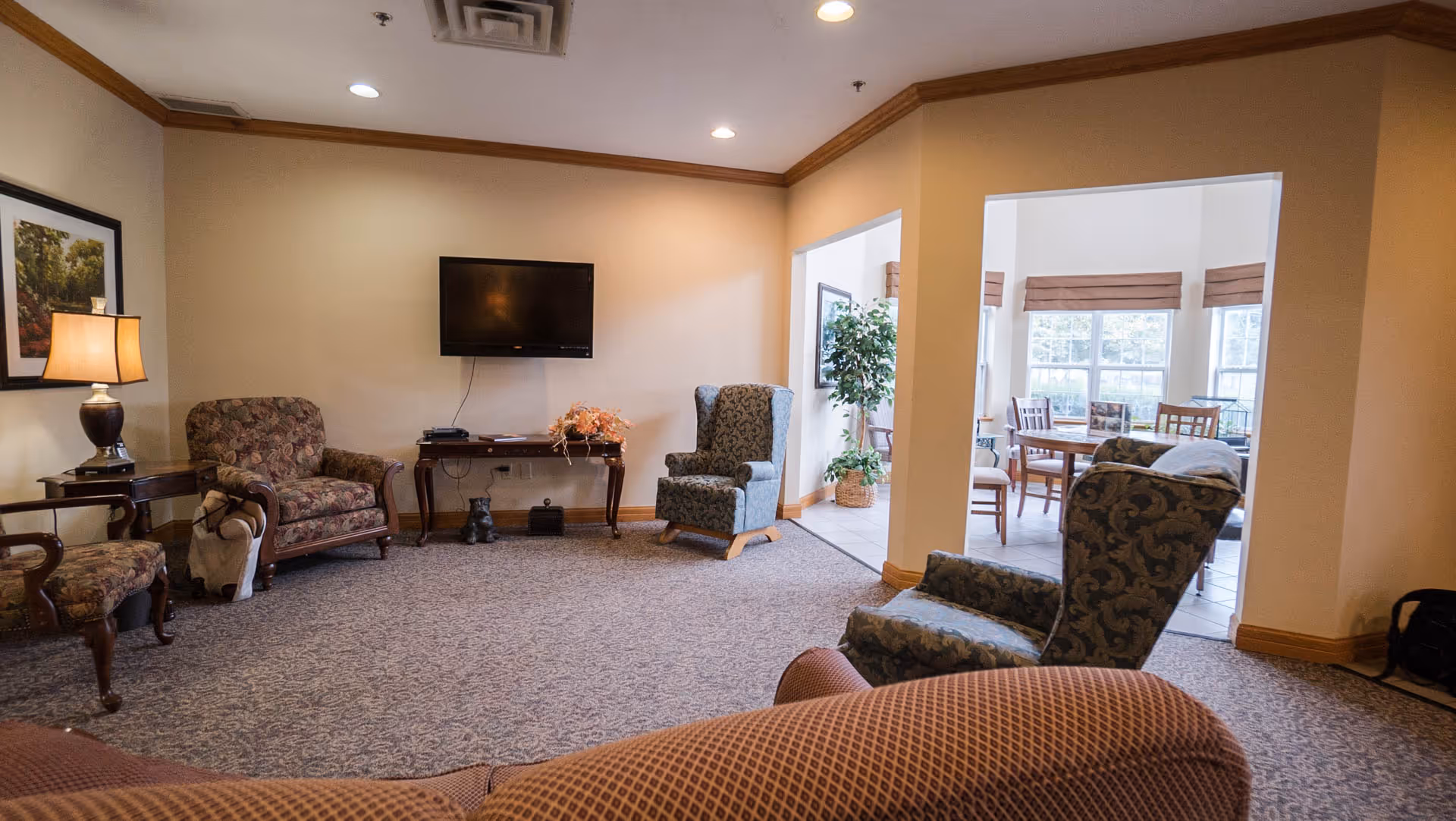 Communal living room with upholstered armchairs, a wall-mounted TV, side tables, and a view into a sunlit dining area.