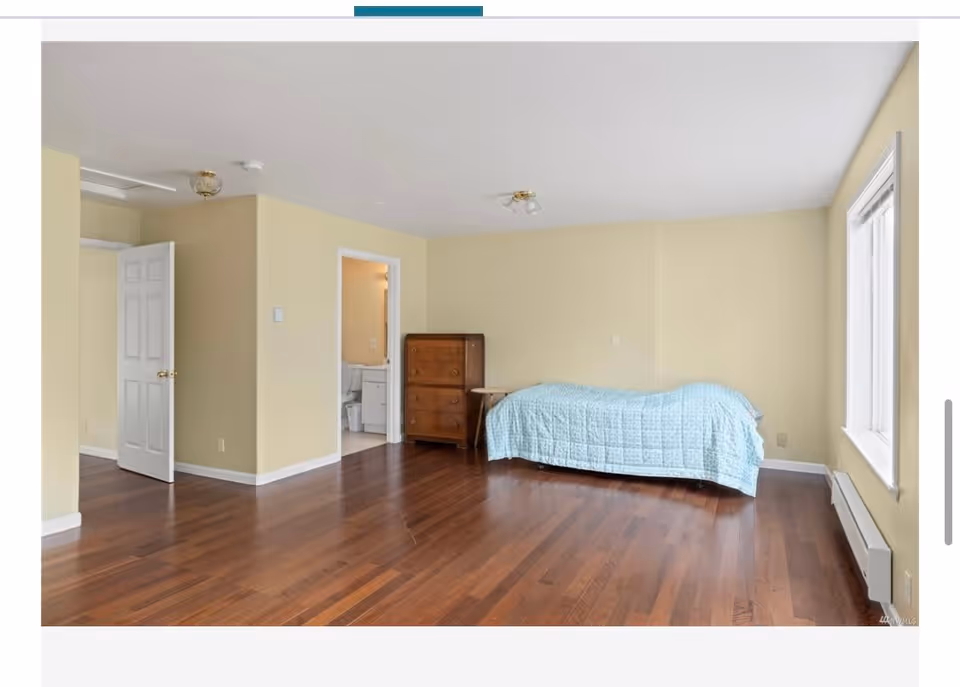 A spacious bedroom with wooden flooring, a single bed covered with a light blue quilt, a wooden dresser, and a small side table. There is a large window on the right wall allowing natural light to enter. An open door leads to an adjacent bathroom.