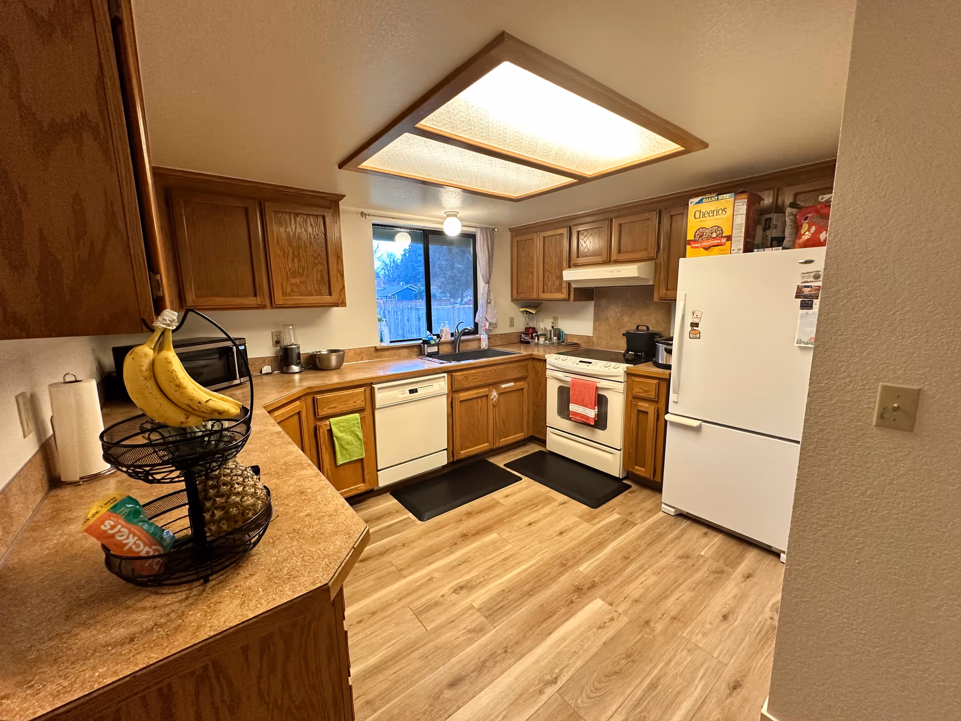 Compact kitchen with wooden cabinets, fridge, stove, sink, and fruit basket.