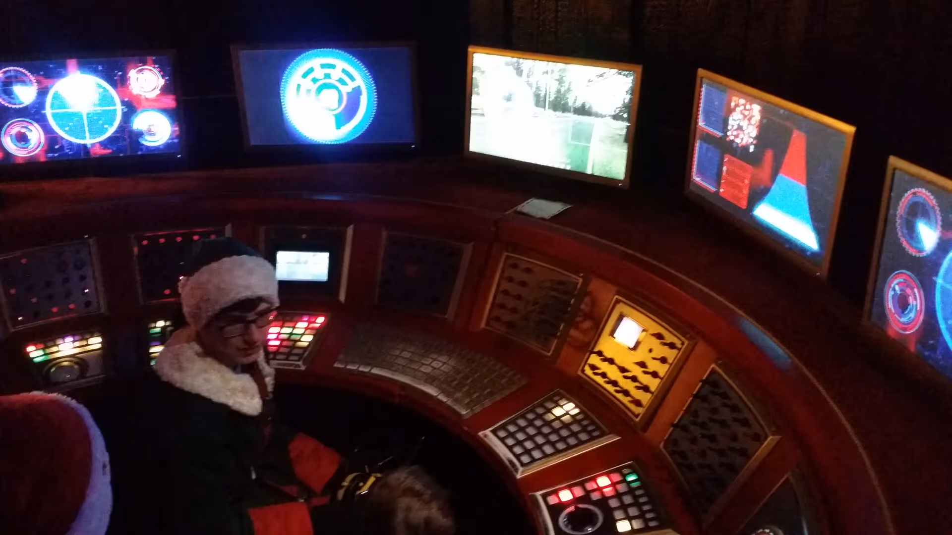 A person wearing a fur-lined coat sits at a curved control desk with multiple screens.