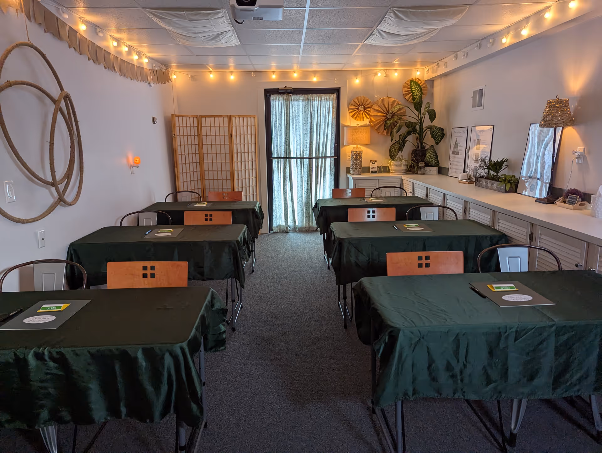 A multipurpose interior room with six green-covered tables, wooden chairs, string lights, and a cabinet counter.