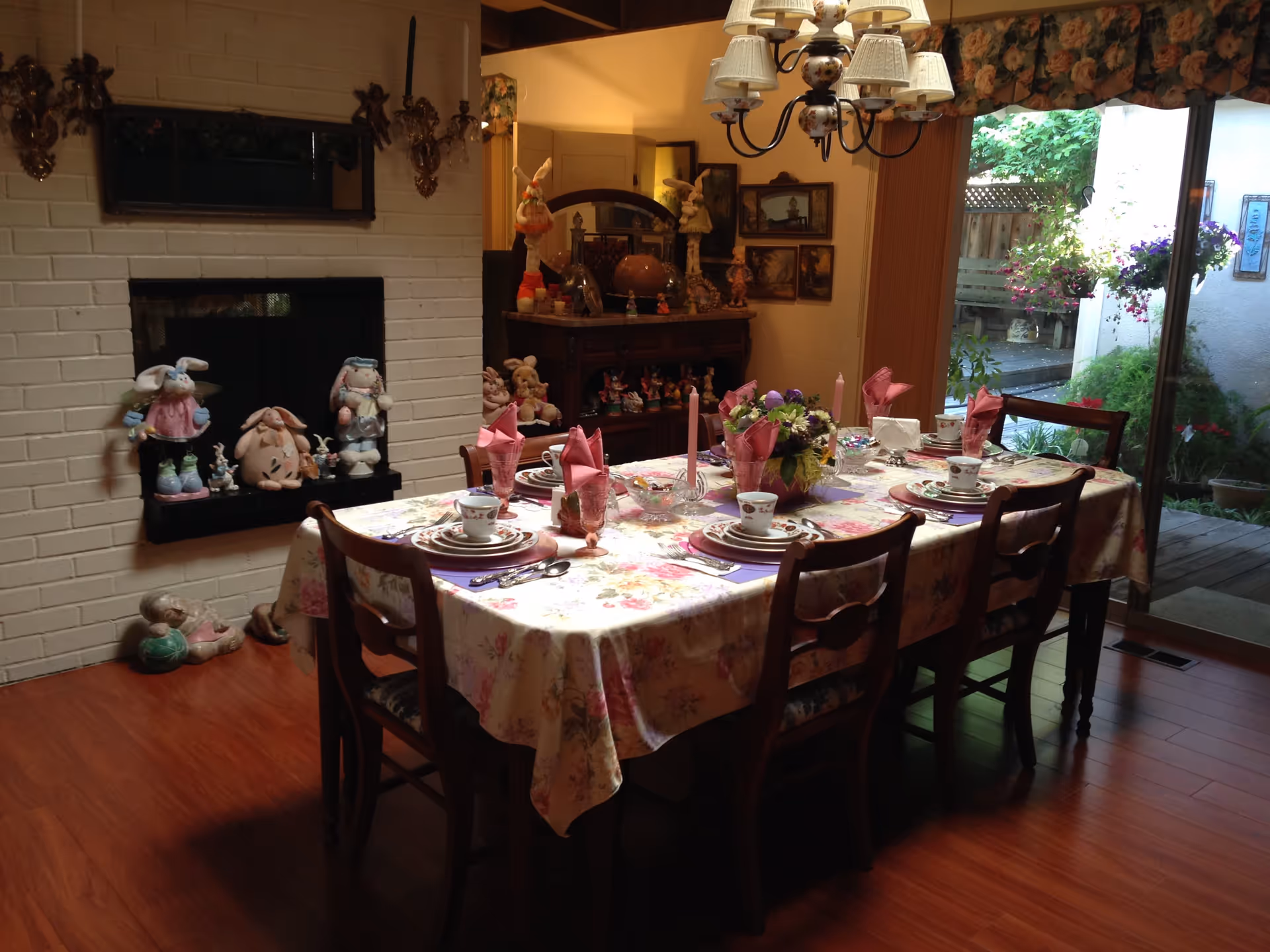 A dining room with a wooden table covered by a floral tablecloth set for six people with plates, cups, pink folded napkins, and candles. The room features a white brick fireplace decorated with bunny figurines, a wooden sideboard with more decorations, a chandelier with floral lampshades, and a large sliding glass door leading to a garden area outside.