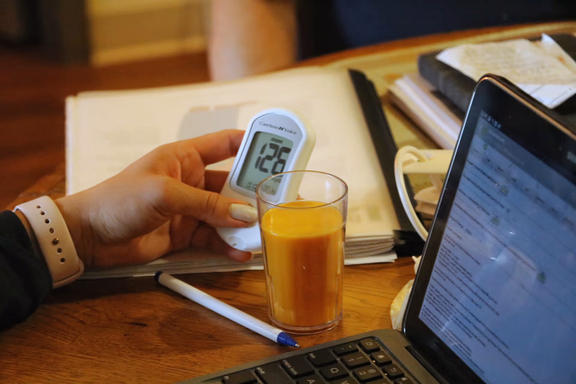A hand holds a glucose meter beside a glass of juice on a desk.