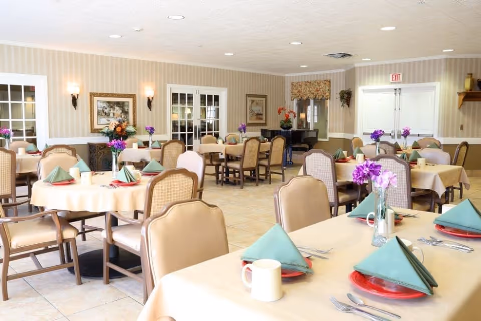 A bright and neatly arranged dining room with multiple tables covered in beige tablecloths, each set with green folded napkins, red plates, white mugs, and silverware. Purple flowers in glass vases decorate the tables. The room has beige striped wallpaper, framed artwork on the walls, and a piano in the corner near a window with floral curtains. There are several cushioned chairs around each table.