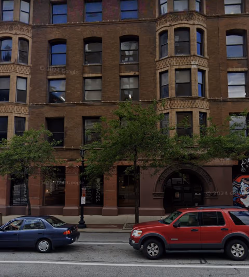 Brick city building facade with arched entrance, parked cars, and green trees along sidewalk.