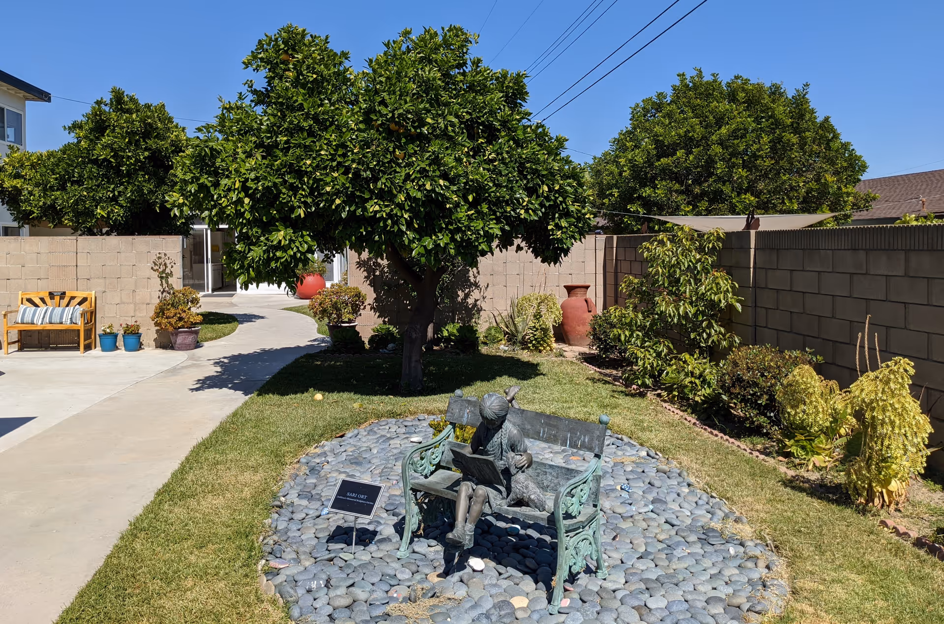 Outdoor garden area with a statue of a girl sitting on a bench reading a book, surrounded by smooth stones. There is a tree with green foliage in the background, along with various potted plants and shrubs. A wooden bench with a striped cushion is placed on a concrete path leading to a building entrance under a clear blue sky.