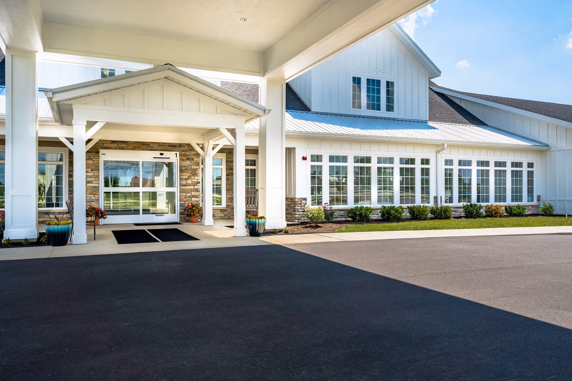 Exterior view of the entrance to a senior living facility named Sugar Fork Crossing, featuring a covered drop-off area with white pillars, large windows, stone accents, and a paved driveway.