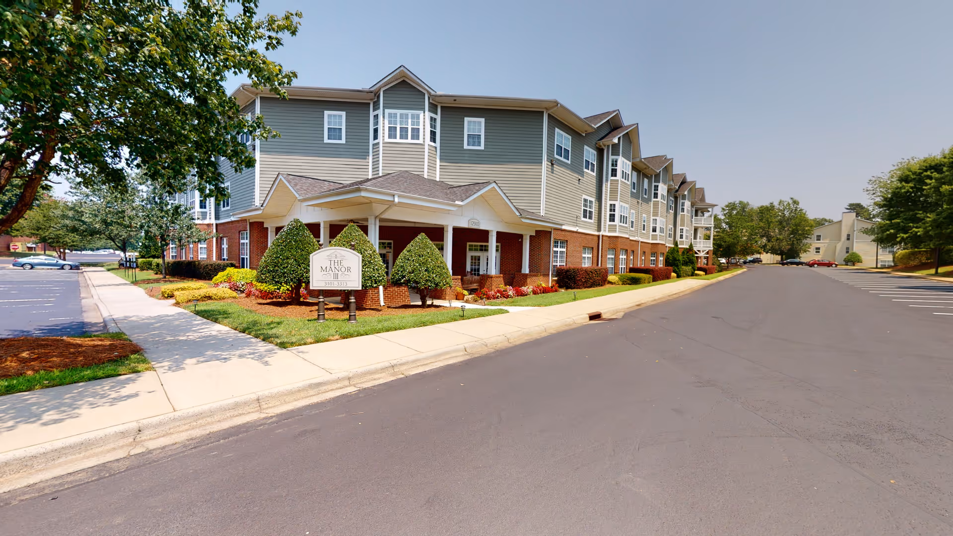 Exterior view of The Manor senior living facility showing a multi-story building with gray and brick facade, a covered entrance, neatly trimmed bushes, and a large parking lot under a clear sky.