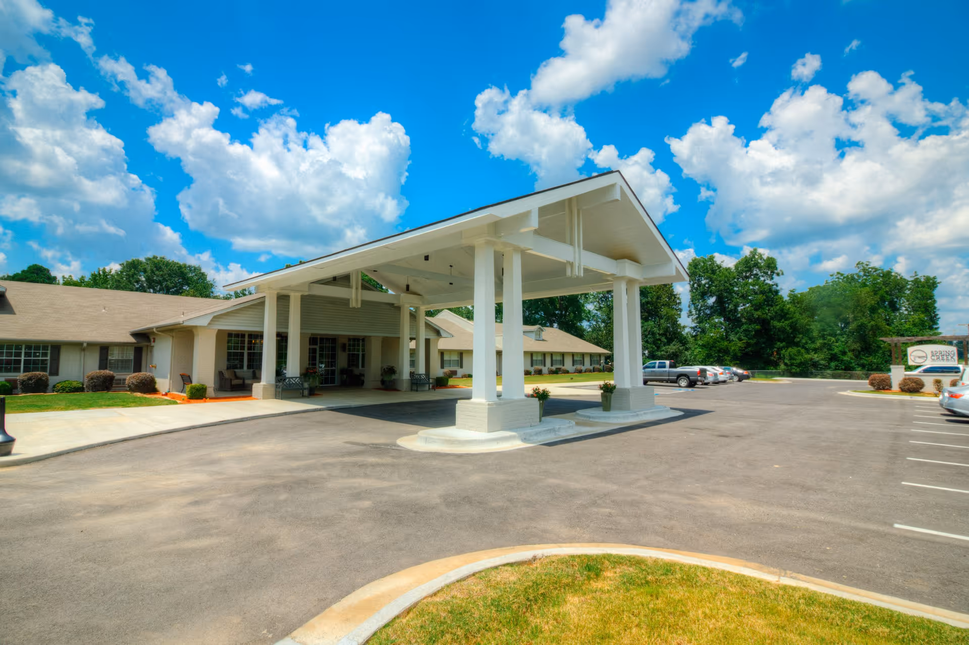 Exterior view of Spring Creek Health & Rehab facility showing a covered entrance with white pillars, a parking lot with several cars, green trees in the background, and a bright blue sky with scattered clouds.