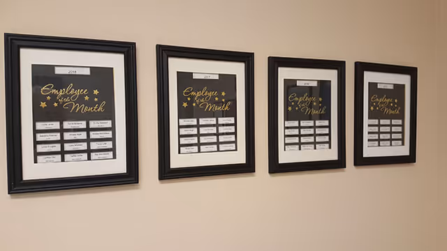Four framed Employee of the Month boards hung along a beige wall in a facility.