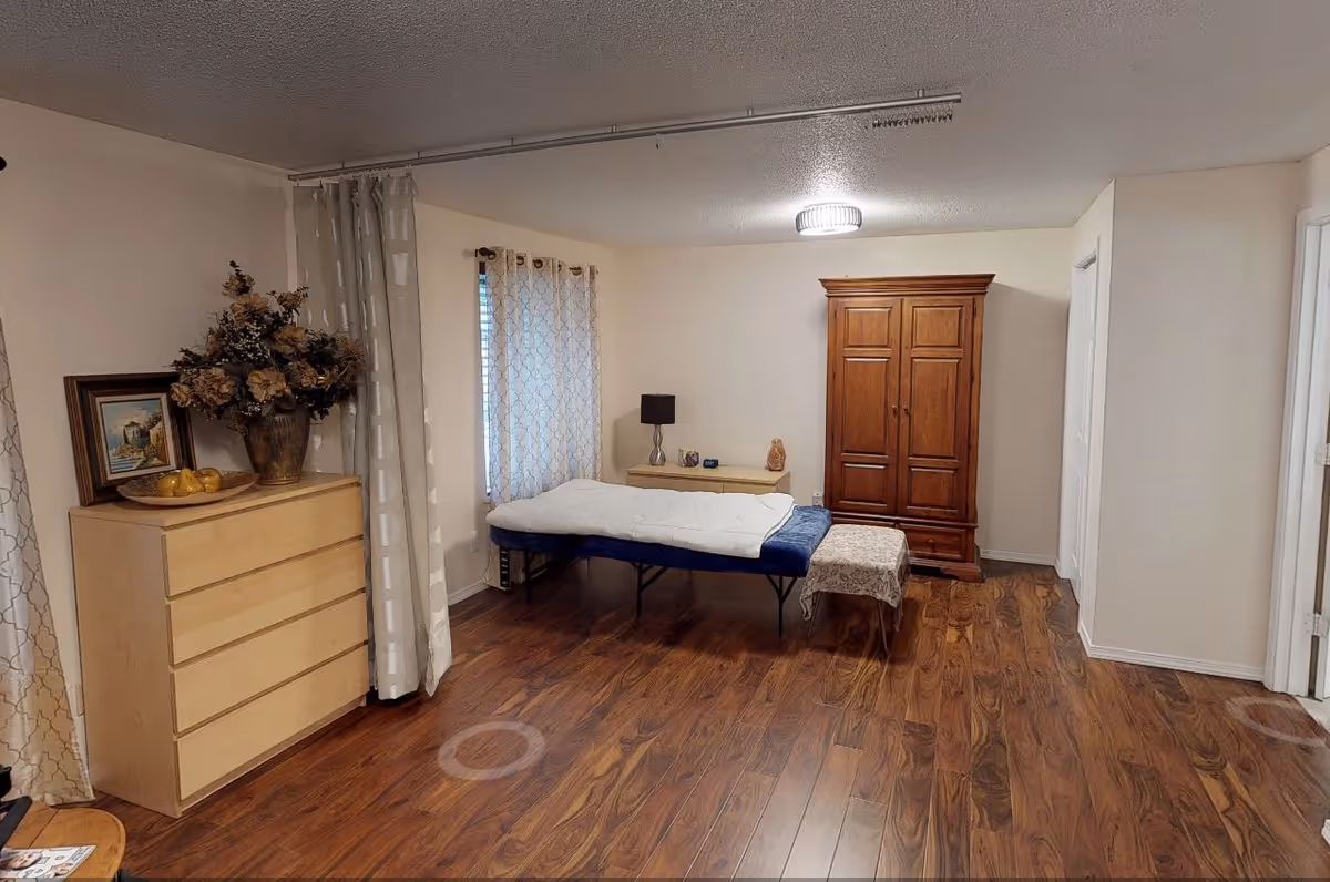 A bedroom in a senior living facility featuring a bed, a wooden dresser, a tall armoire, and curtains by the window.