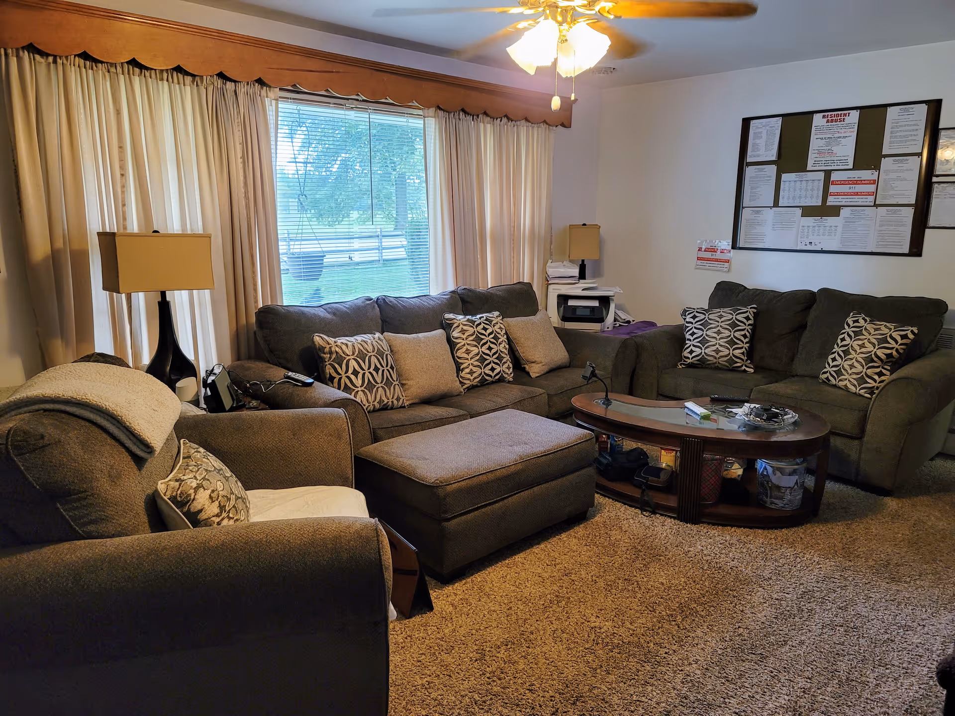A cozy living room with a large window covered by beige curtains. The room features a brown sofa set with patterned and plain cushions, a matching ottoman, and a wooden coffee table with various items on it. There are two table lamps on either side of the sofas, a ceiling fan with lights, and a bulletin board with notices on the wall.