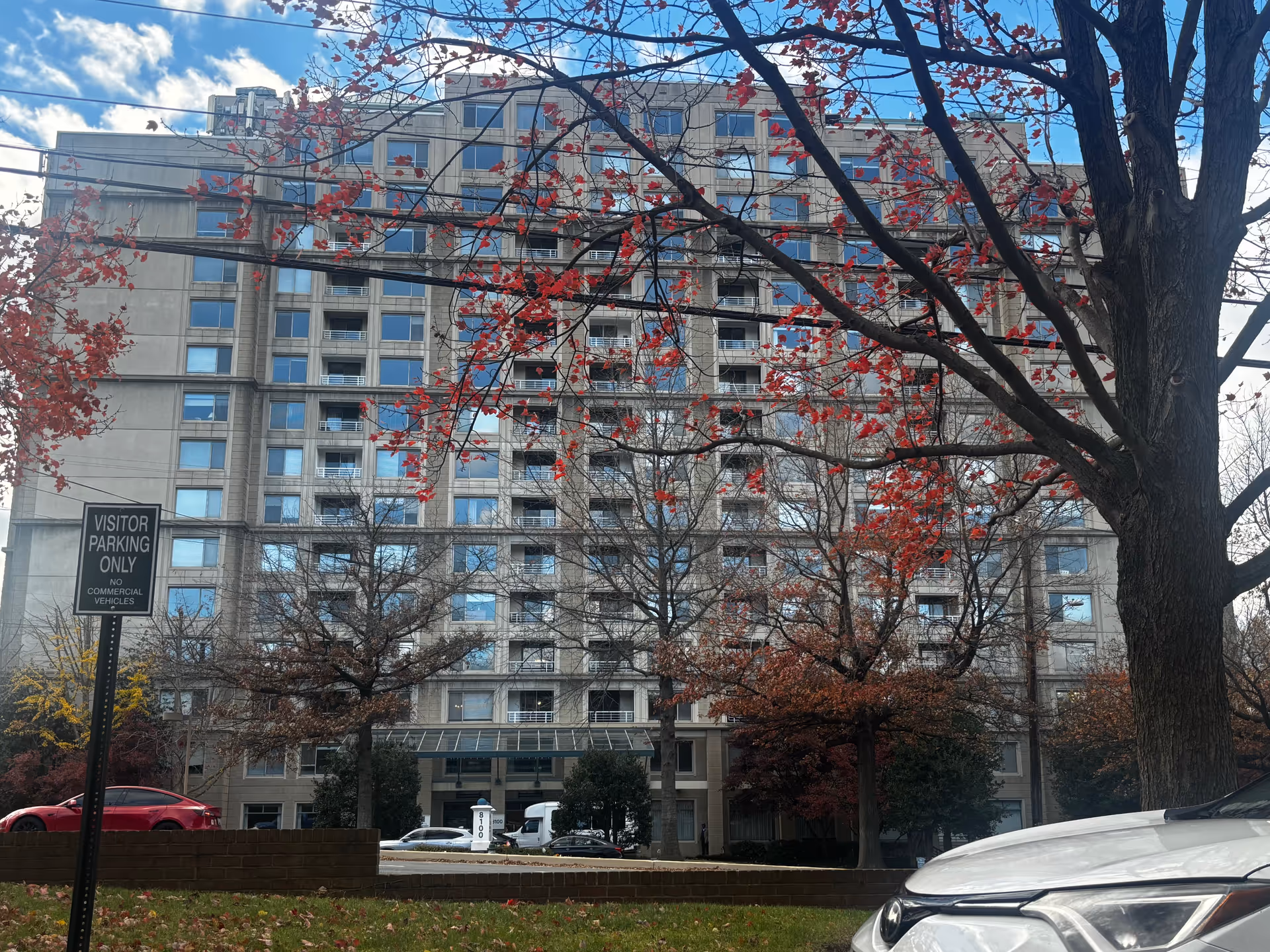 Multi-story residential building front with red-leaved trees, parked cars, and a visitor parking sign.
